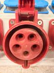 Red round CEE socket with hinged lid and five contact holes on a white box, blue sockets above