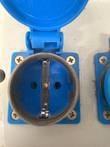 Blue European-style outlet with lid open, black seal ring and metal earth clips on a white box