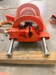 Orange grooving machine from the clamp side: V-jaw vise, mesh motor guard and chrome guide rods resting on a wood beam