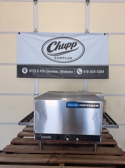Lincoln Impinger conveyor oven on legs on a wooden pallet, conveyor belt out both sides, control knobs on front