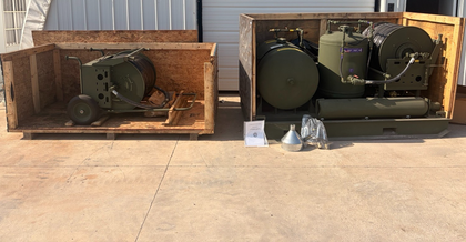 Two open crates: wheeled hose reel at left, green tanks and reel skid at right, with funnel and manual